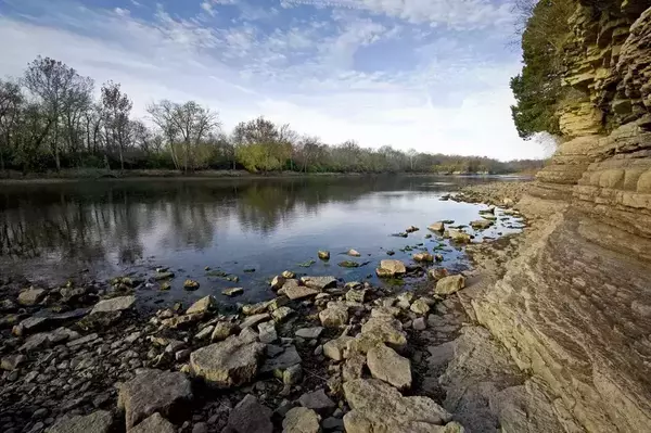 A wide, calm river reflects bare trees and a blue sky with cirrus clouds. A rocky bank with scattered stones lines the left shore. On the right, layered tan rock cliffs with hints of green foliage rise steeply from the water.