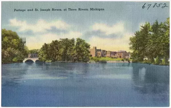 View of Portage and St. Joseph Rivers in Three Rivers, Michigan, with a stone arch bridge, trees, and buildings along the banks.